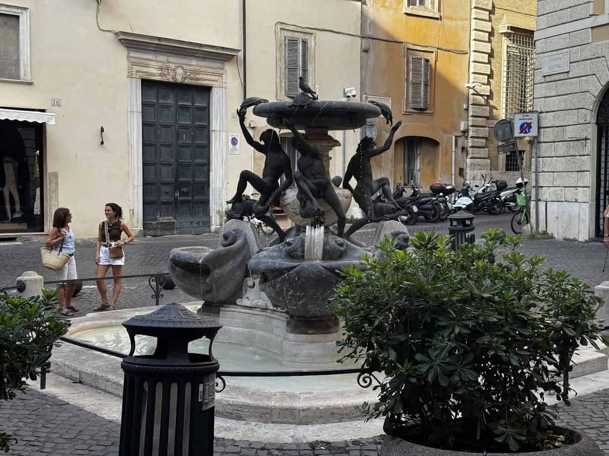 A fountain with dolphin and human statues in a small square, perfect for a stop on your Rome with Kids adventure, with people walking nearby and buildings behind.