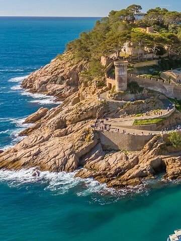 Aerial view of a seaside castle on a rocky hill beside a sandy beach with boats in turquoise water.