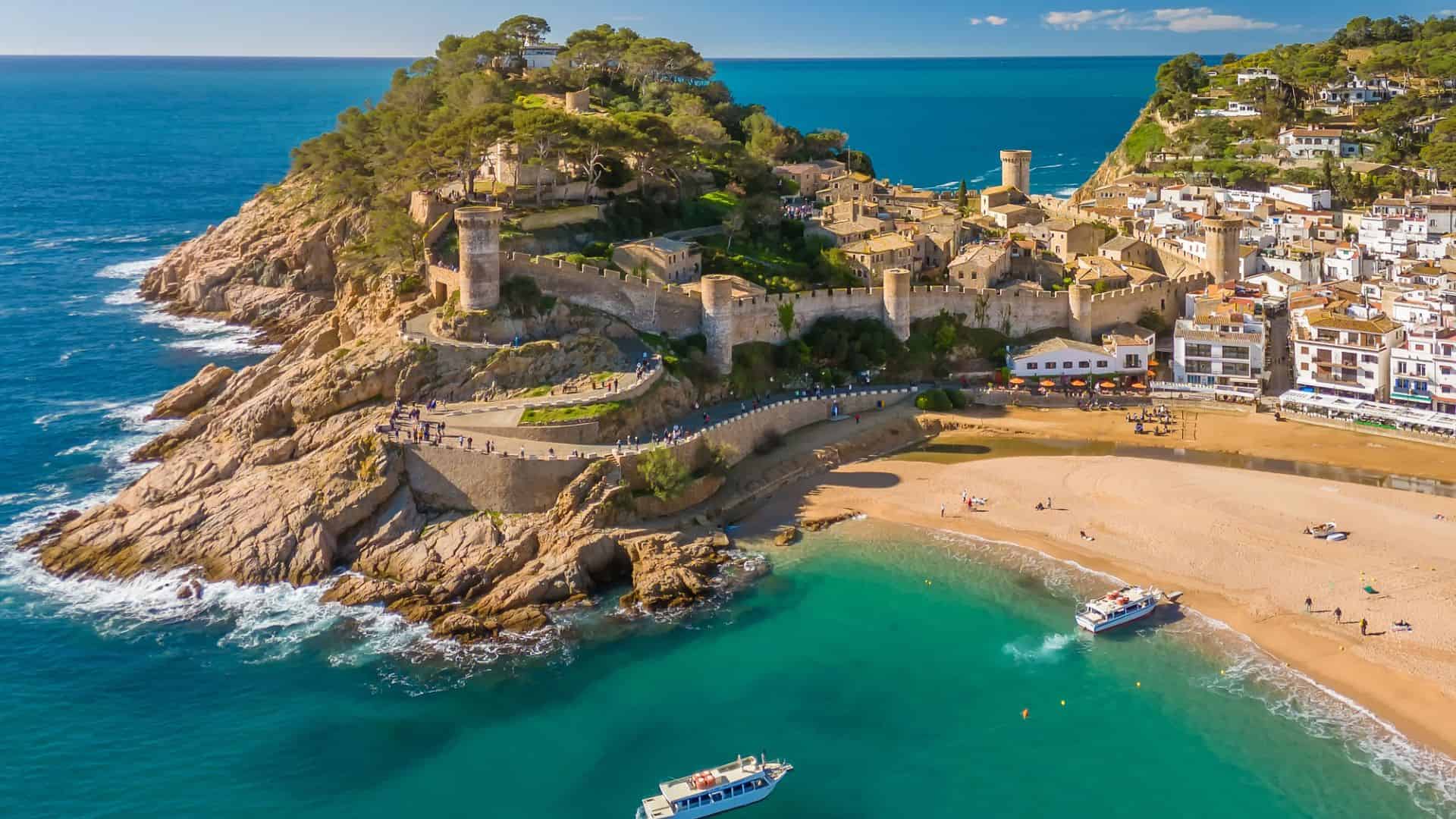 Aerial view of a seaside castle on a rocky hill beside a sandy beach with boats in turquoise water.