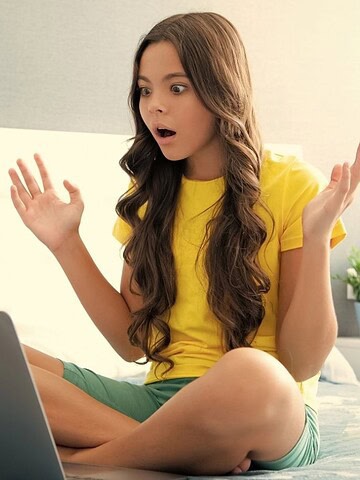 A surprised girl sits cross-legged on a bed, looking at a laptop with her hands raised.