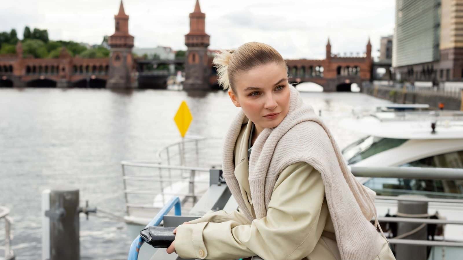 Woman in light coat leaning on a railing by a river, with a city bridge and buildings in the background.