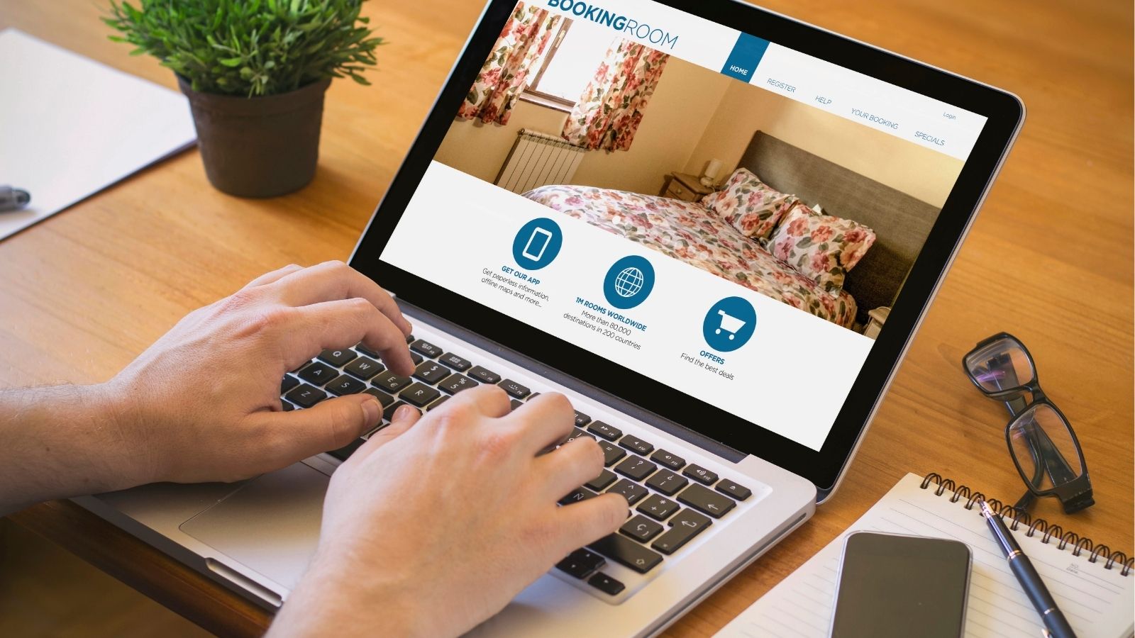 Person using a laptop to book a hotel room online, with a notebook, pen, and plant on the desk.