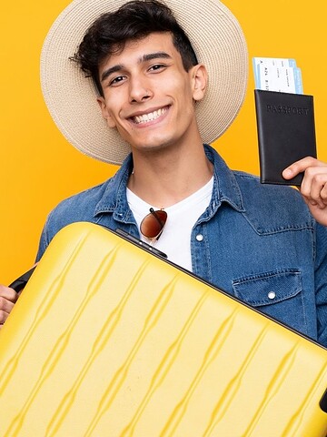 Smiling person with a hat holds a yellow suitcase and a passport with boarding passes, standing by a yellow background.