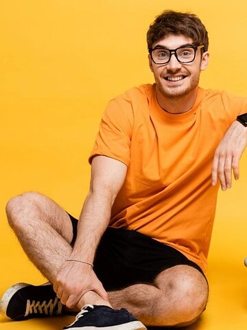 Smiling man in orange shirt and glasses sits next to a yellow suitcase against a yellow background.