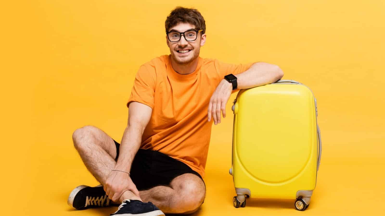Smiling man in orange shirt and glasses sits next to a yellow suitcase against a yellow background.