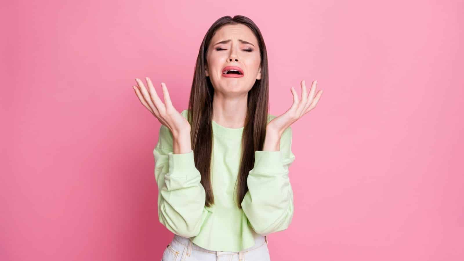 A young woman with long hair looks upset and raises her hands while crying against a pink background.