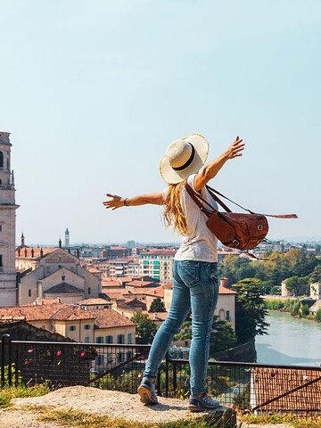 Woman with arms outstretched overlooking a scenic city with historic buildings and a river on a sunny day.