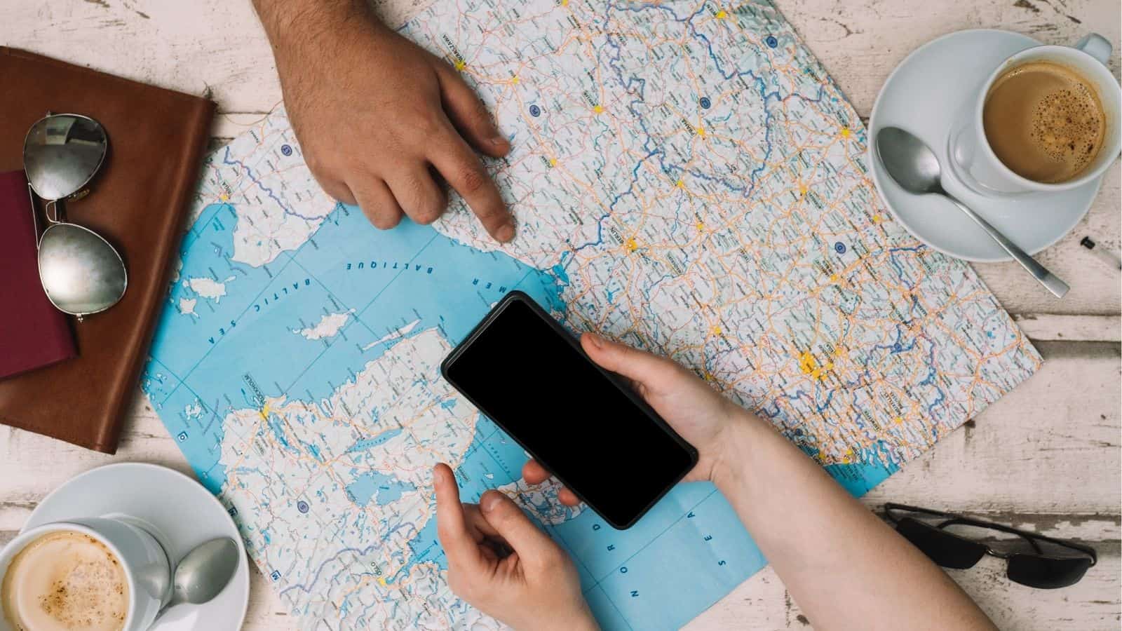 Two people planning a trip with a map, smartphone, coffee cups, sunglasses, and a passport on a table.