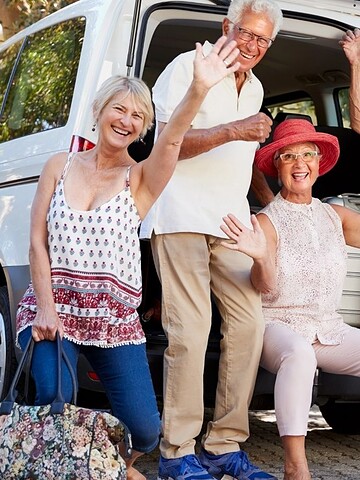 Five smiling seniors waving by a car with open trunk, ready for a trip, in a sunny driveway.