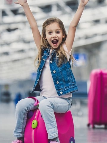 Smiling girl with arms raised sits on pink suitcase; two adults sit in the background at an airport.