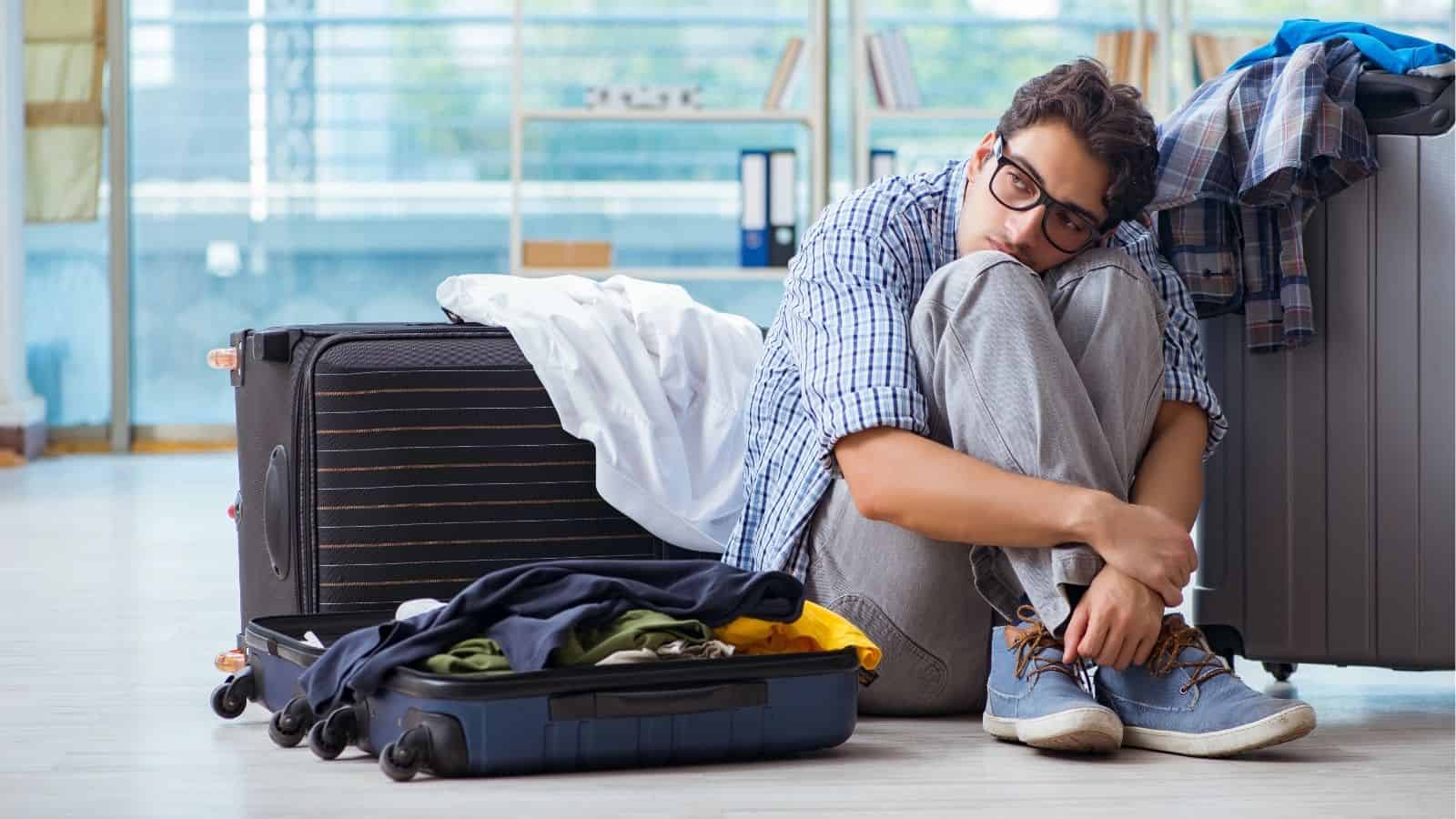 A young man sits on the floor looking stressed, surrounded by open suitcases and scattered clothes.