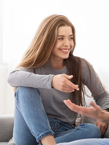 A smiling woman and man sit on a couch, talking and gesturing to each other in a bright living room.