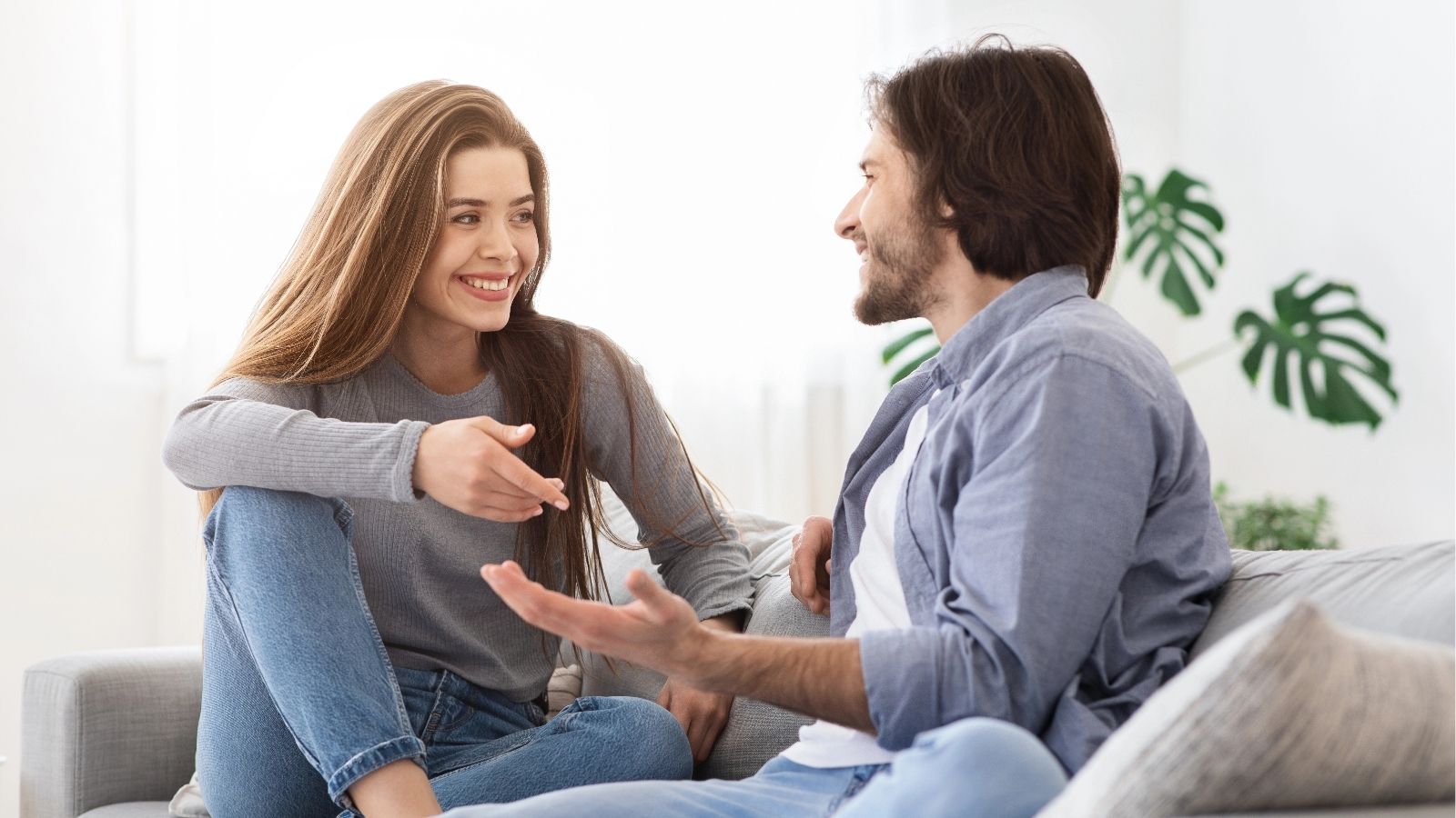 A smiling woman and man sit on a couch, talking and gesturing to each other in a bright living room.