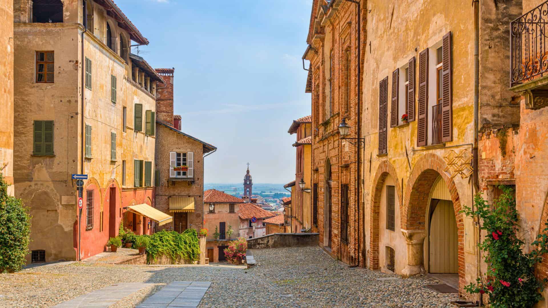 A narrow cobblestone street lined with old brick buildings, leading to a distant church tower under a blue sky.