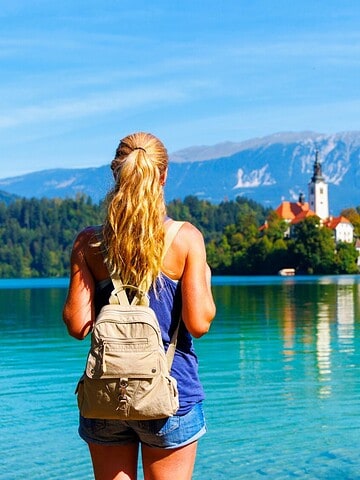 A woman with a backpack looks at a lake and distant church, with mountains in the background.
