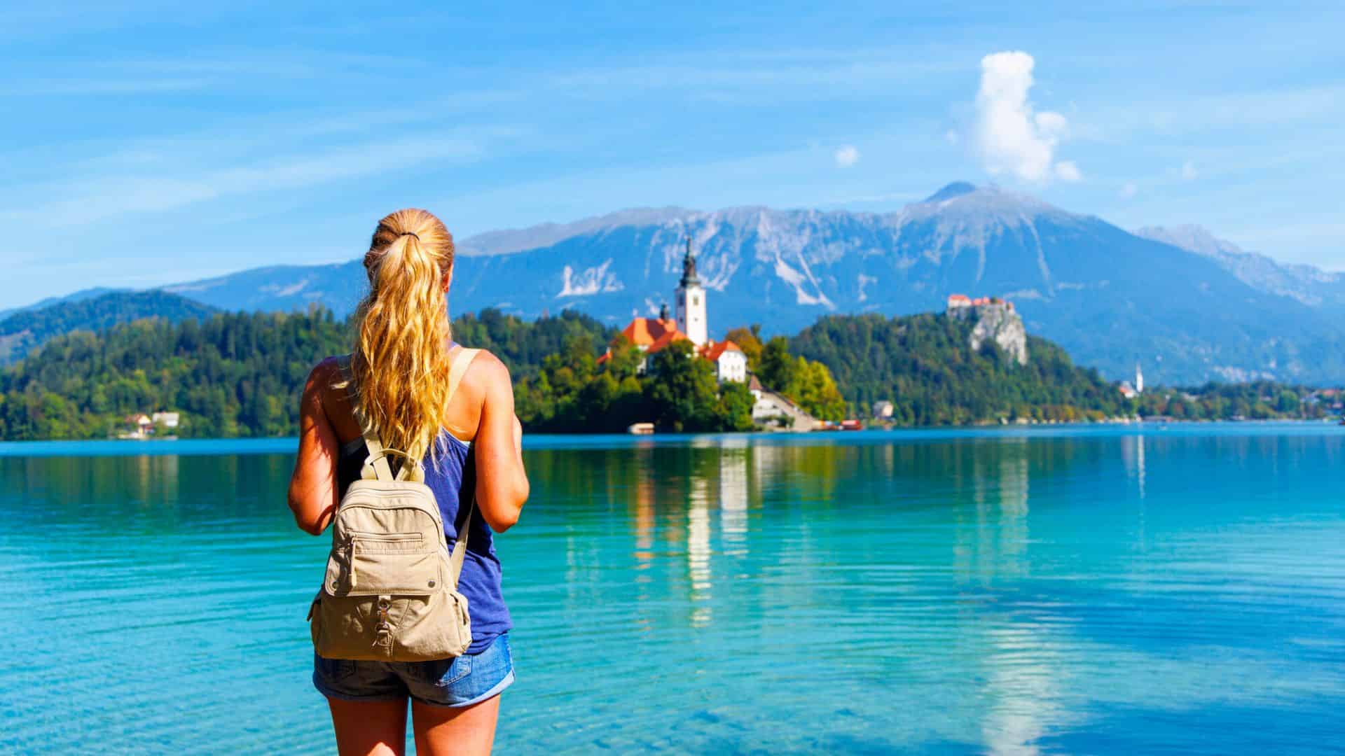 A woman with a backpack looks at a lake and distant church, with mountains in the background.