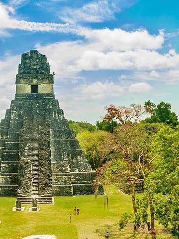Ancient stone pyramid temple surrounded by lush greenery under a bright blue sky with white clouds.