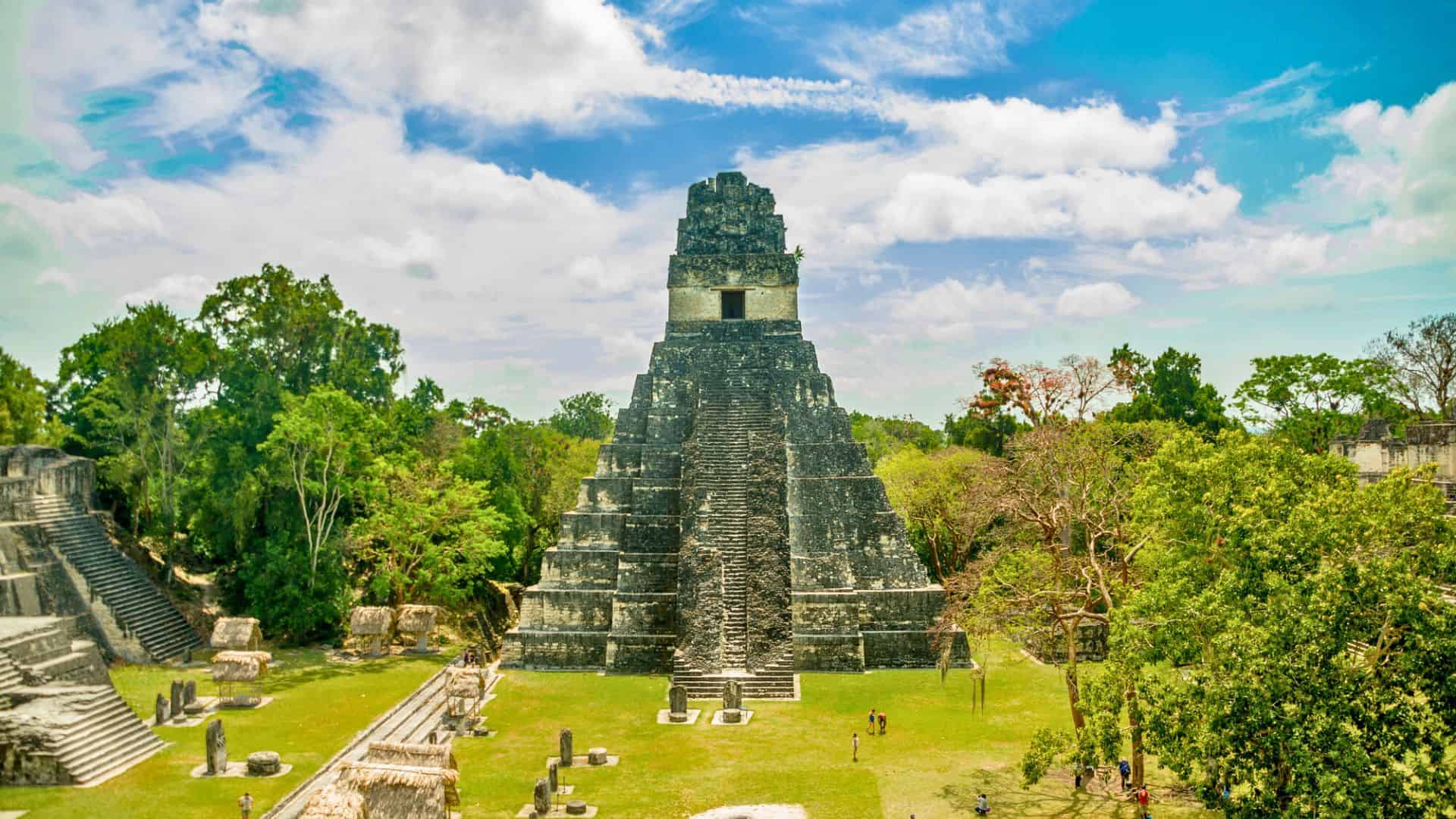 Ancient stone pyramid temple surrounded by lush greenery under a bright blue sky with white clouds.