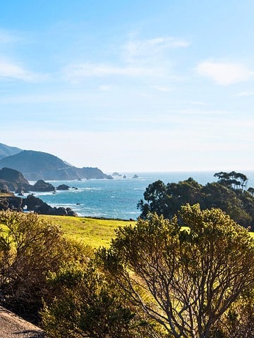 A white car parked on a coastal road with mountains and ocean in the background under a clear blue sky.