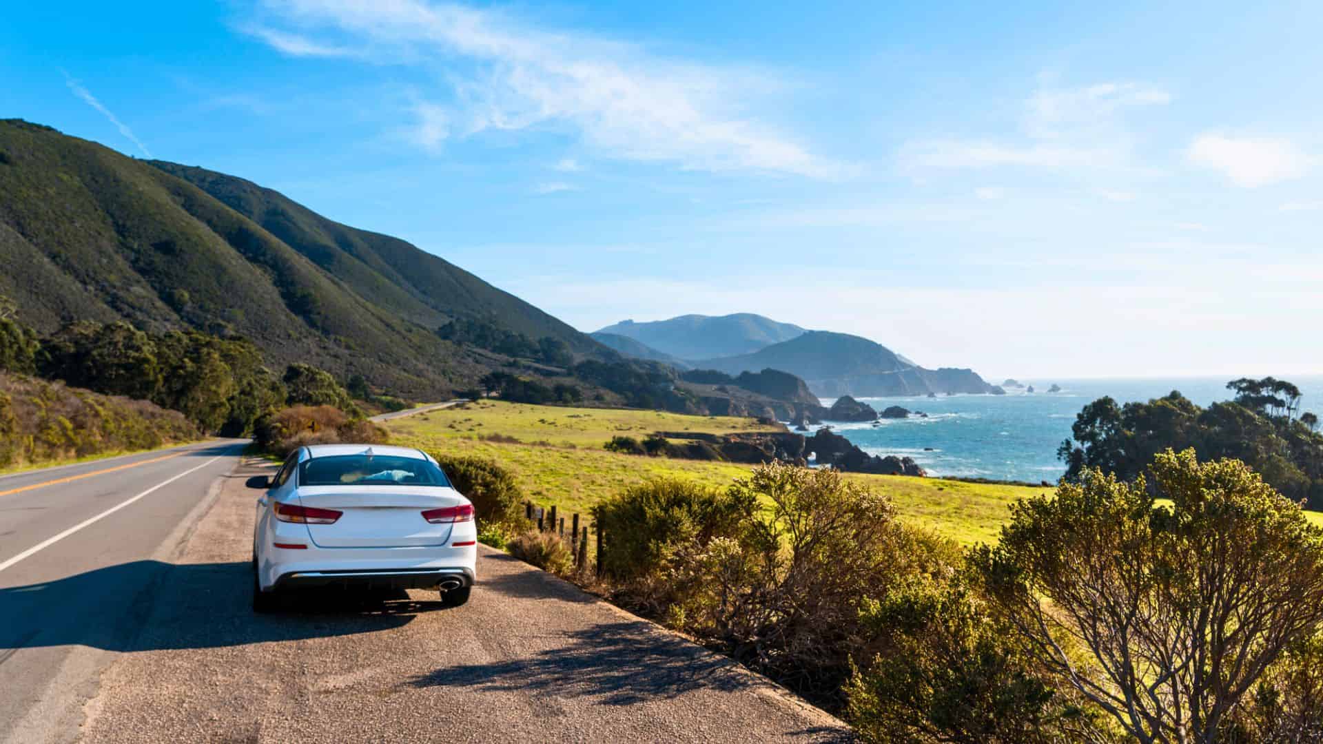 A white car parked on a coastal road with mountains and ocean in the background under a clear blue sky.