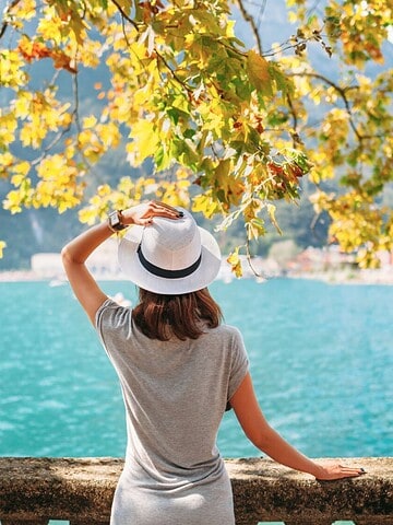 Woman in a hat stands by a lakeside stone railing, facing blue water and mountains under leafy branches.