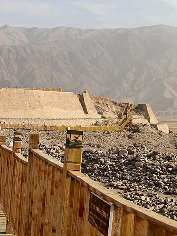 Tourists walk on a wooden bridge toward a large ancient fortress set against a backdrop of rugged mountains.