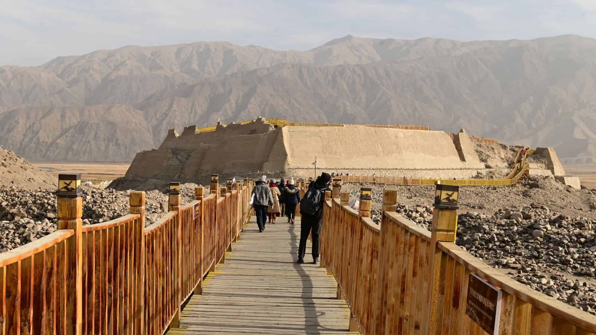 Tourists walk on a wooden bridge toward a large ancient fortress set against a backdrop of rugged mountains.
