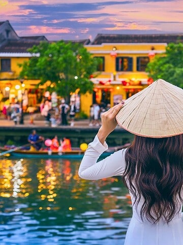 Woman in traditional hat stands by a river, watching boats and colorful buildings at sunset in a lively town.