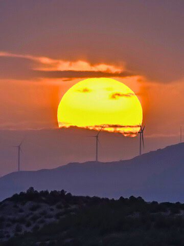 The sun sets behind hills with wind turbines, casting an orange and purple glow over the landscape.