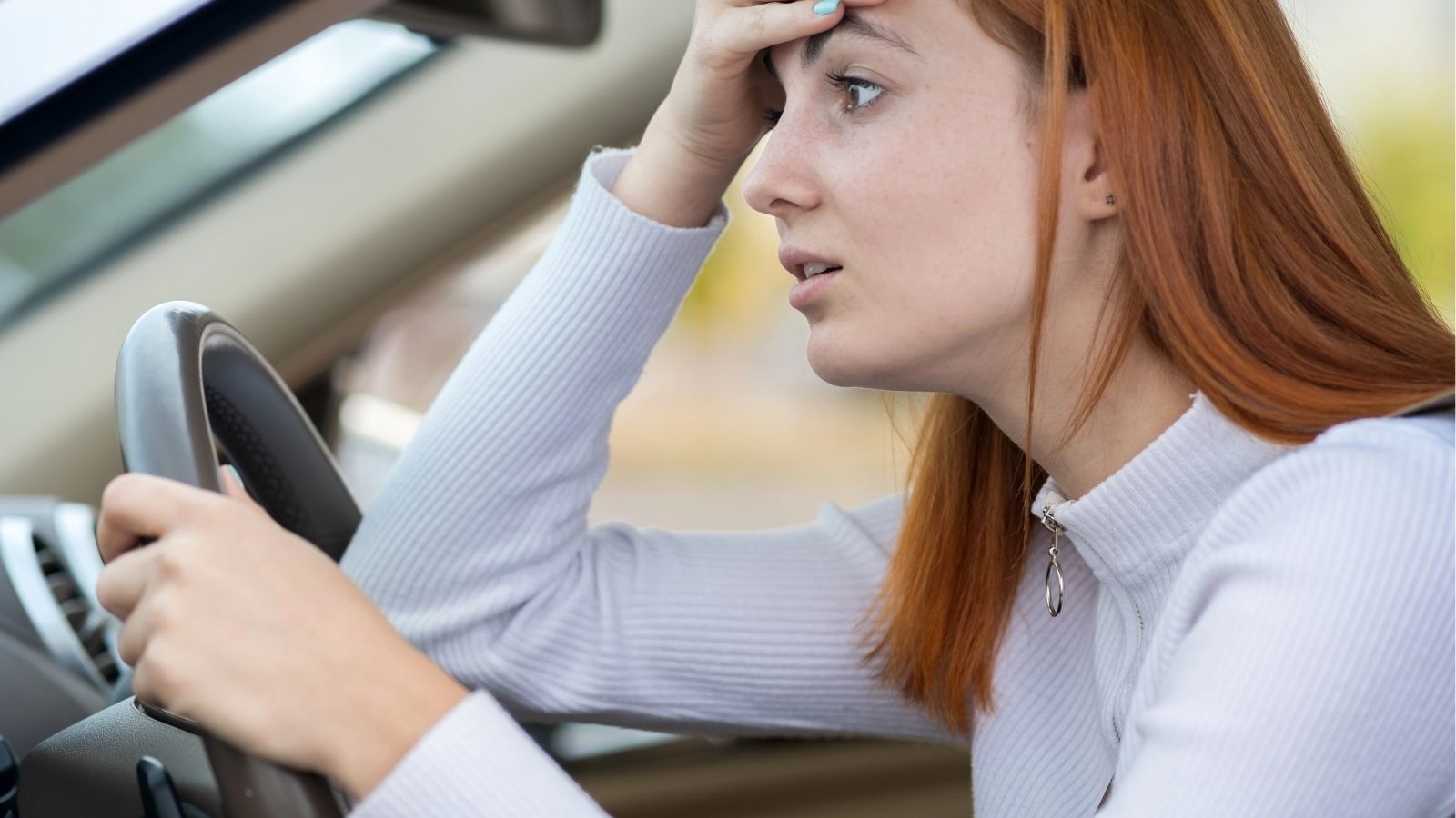 A worried woman with red hair sits in a car, gripping the steering wheel and resting her hand on her forehead, anxious about driving in difficult countries while she travels.