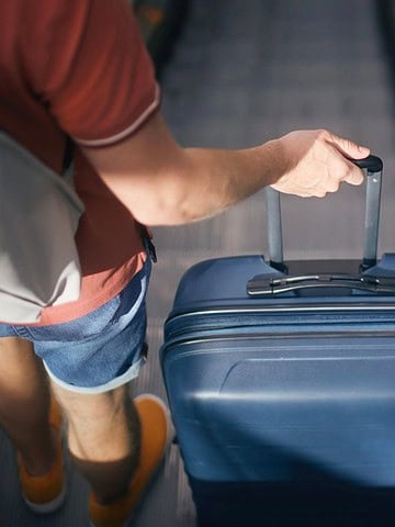 Person in shorts with a backpack pulls a blue suitcase on an escalator, seen from above.