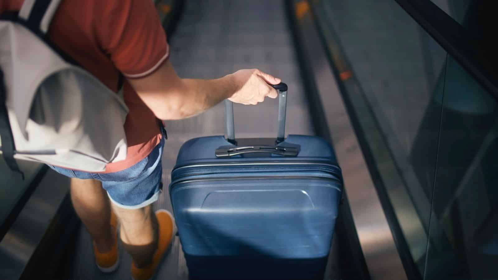 Person in shorts with a backpack pulls a blue suitcase on an escalator, seen from above.