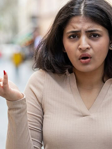 Woman with shoulder-length dark hair shrugging with confused expression outdoors, wearing a beige top.