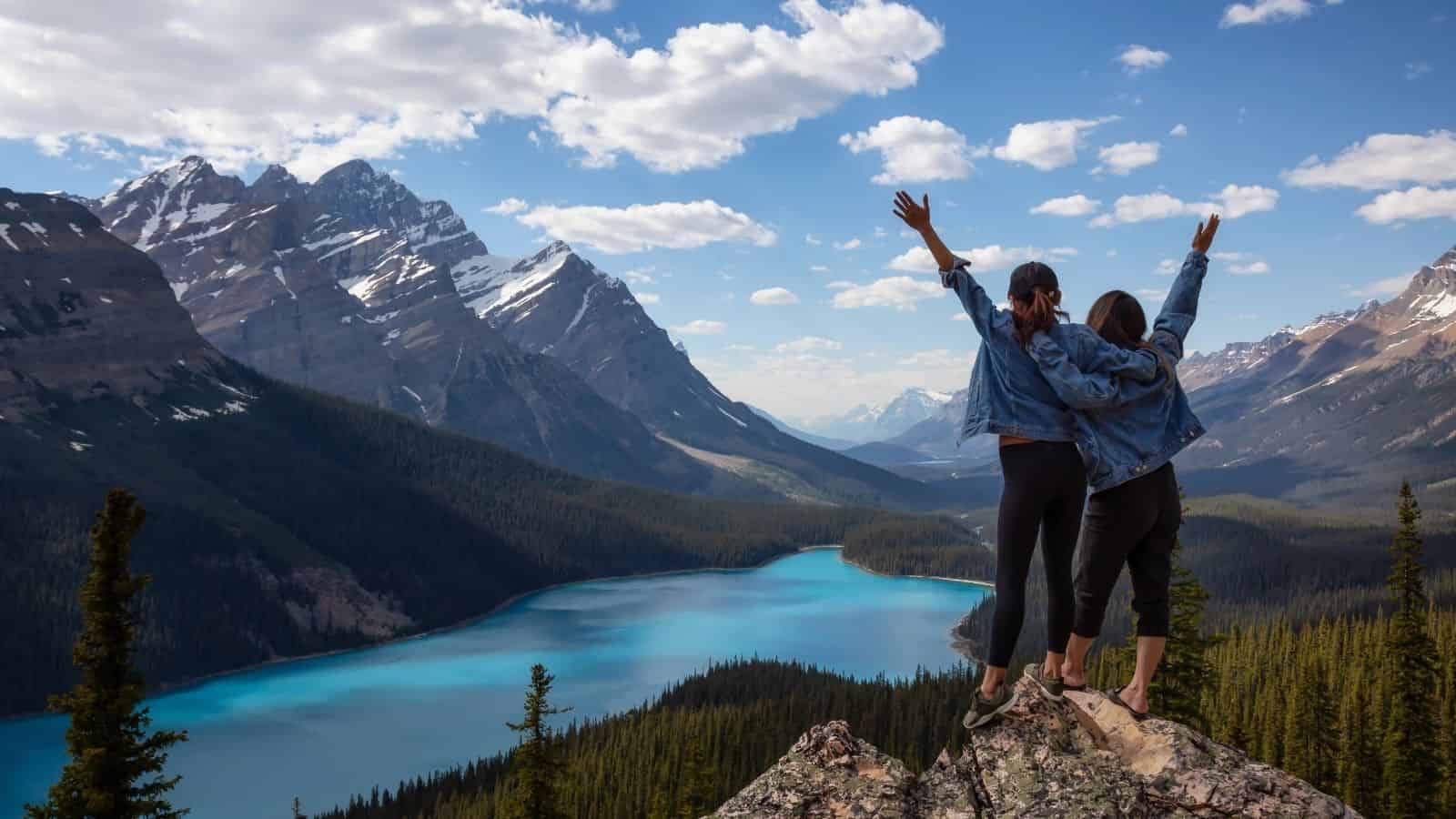 Two people stand on a rocky cliff raising their arms, overlooking a blue lake and snowy mountains&mdash;an unforgettable moment on a Canada vacation.
