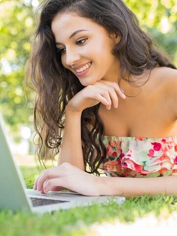 A woman in a floral dress lies on grass, smiling while using a laptop outdoors on a sunny day.