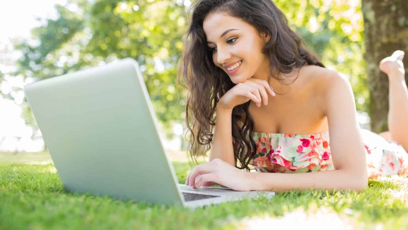 A woman in a floral dress lies on grass, smiling while using a laptop outdoors on a sunny day.
