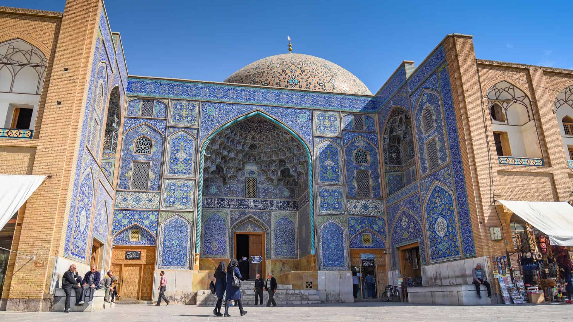 People walk in front of a large mosque with intricate blue tilework and a prominent dome under a clear sky.
