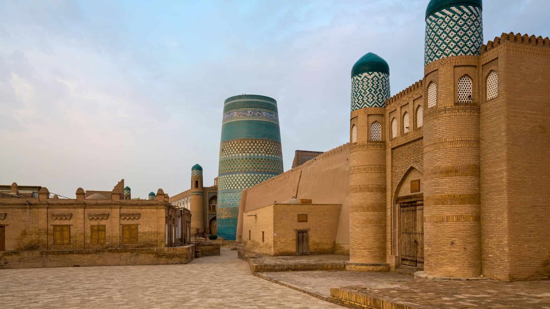 Ancient city walls and turquoise-tiled minarets in Khiva, Uzbekistan, under a partly cloudy sky.
