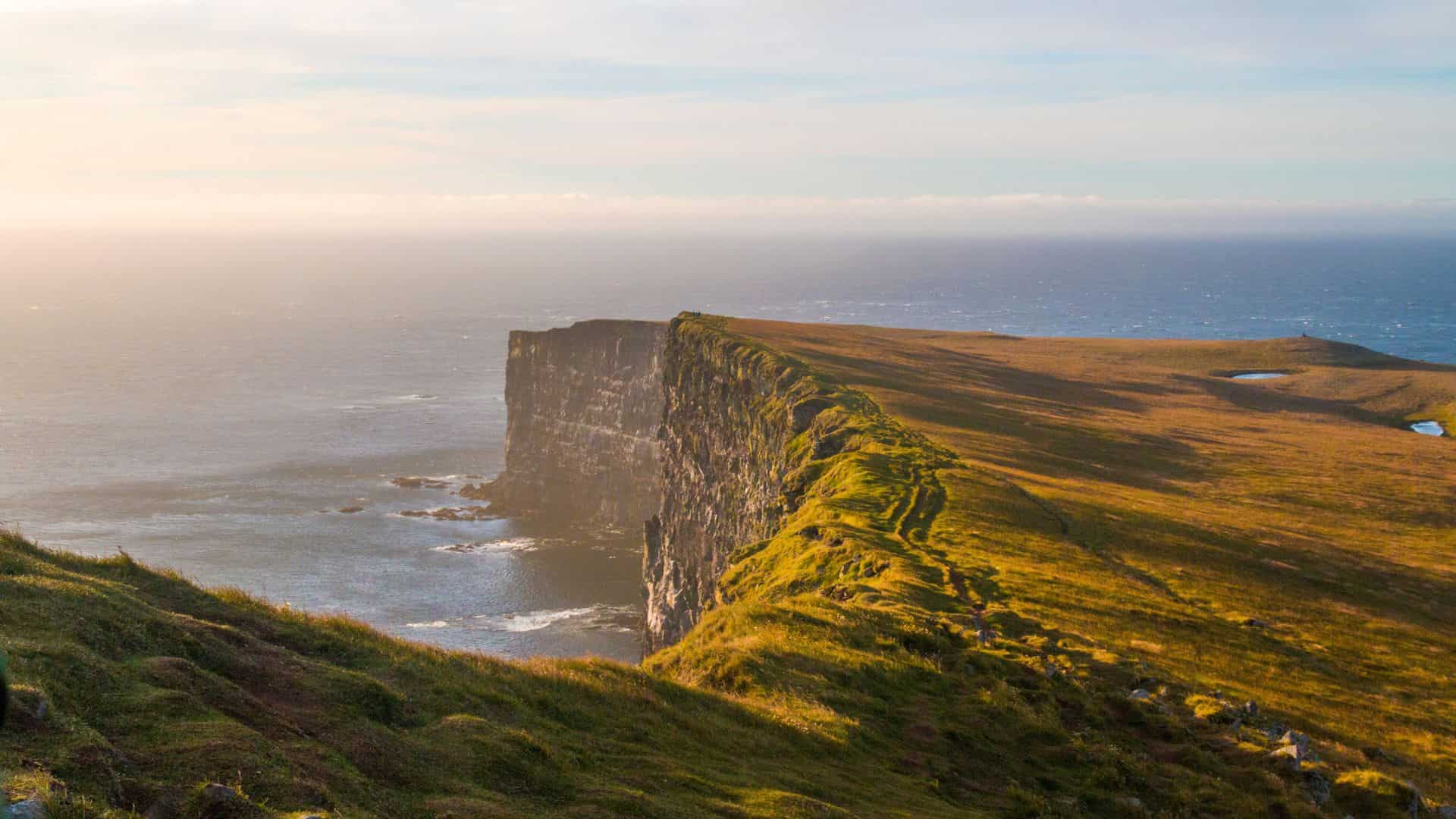 Cliffs covered in green grass overlook the ocean under a hazy sky at sunset.