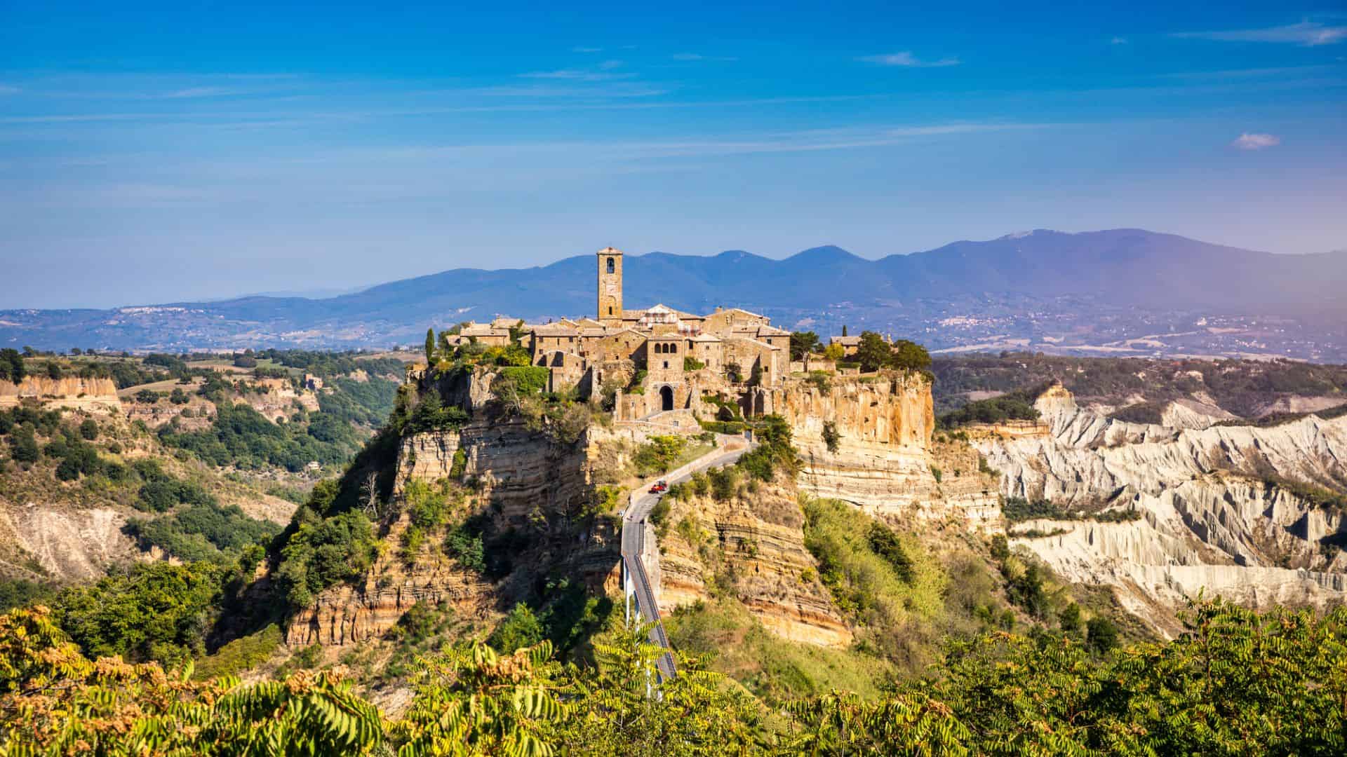 Hilltop medieval village with stone buildings and a tower, surrounded by cliffs and greenery under a blue sky.