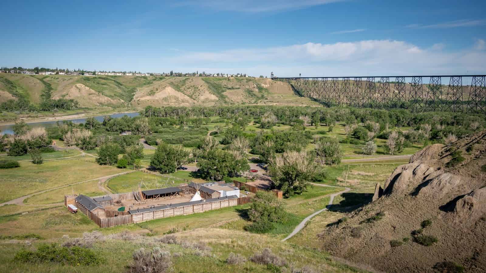 A historic wooden fort sits in a grassy valley with a large railroad bridge in the background.