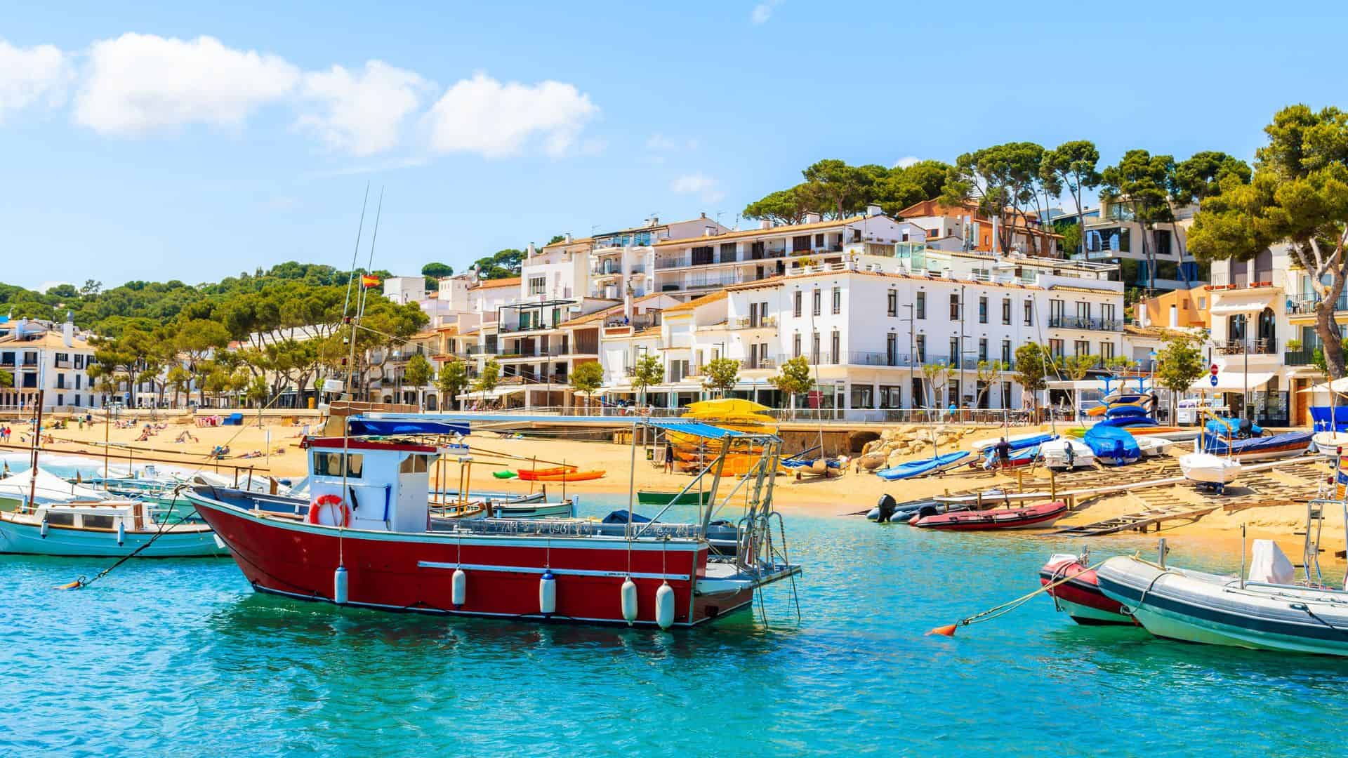 Colorful boats float on blue water with white seaside buildings and sandy beach in the background.