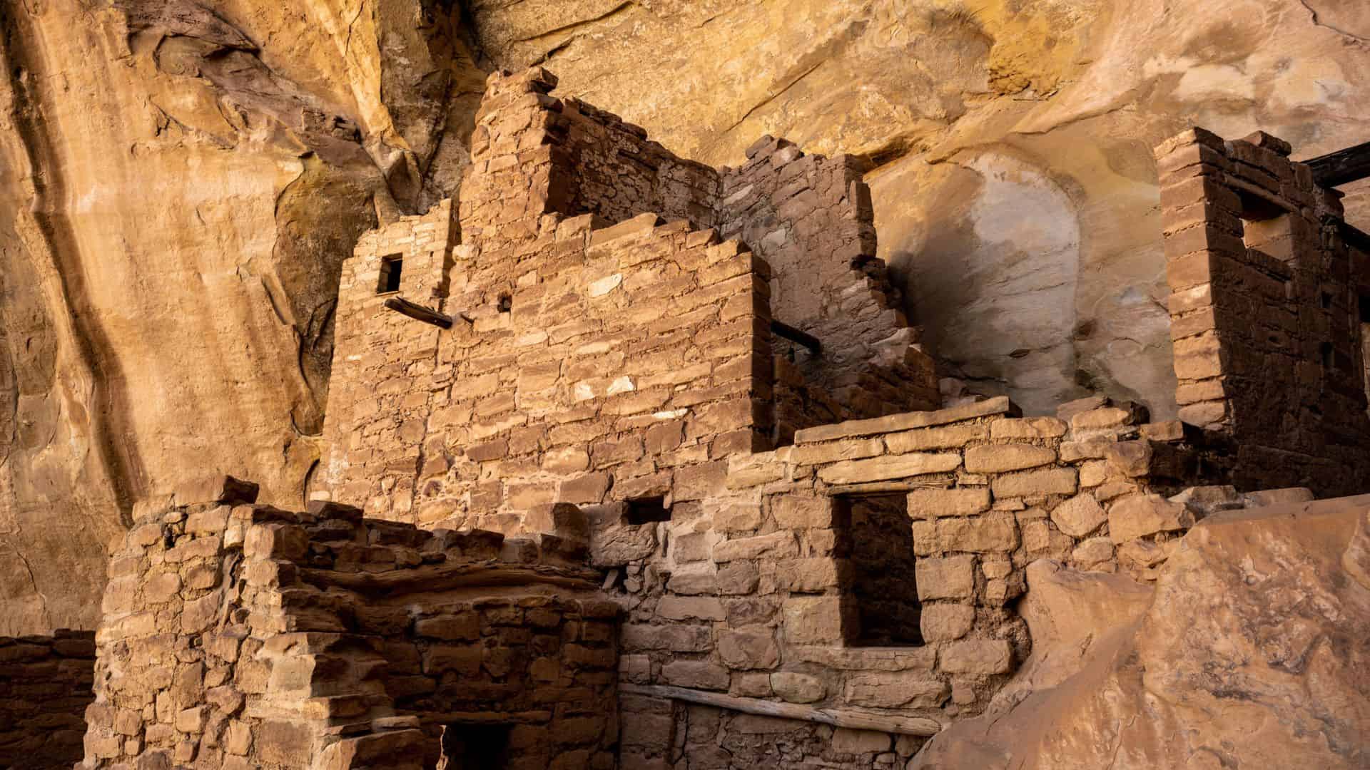 Ancient stone cliff dwellings built into a rocky canyon wall, lit by warm sunlight.