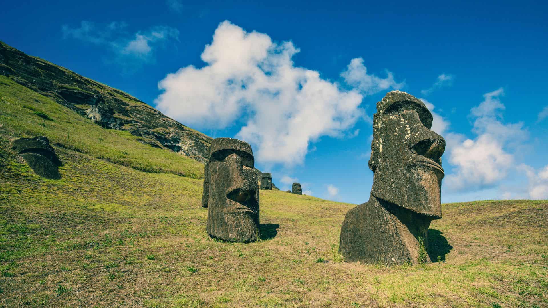 Two large stone Moai statues on a grassy hill under a blue sky with scattered clouds on Easter Island.