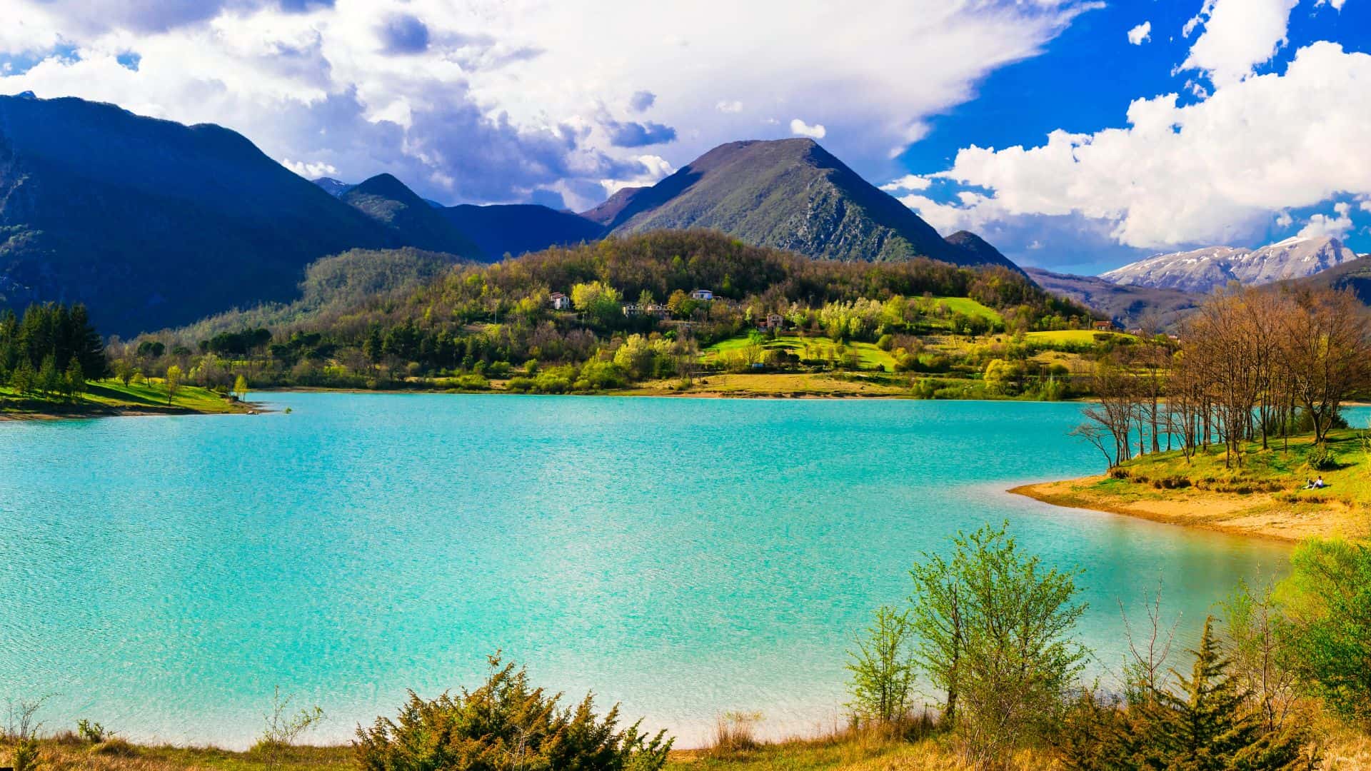 Turquoise lake with green hills, trees, and mountains under a partly cloudy blue sky.