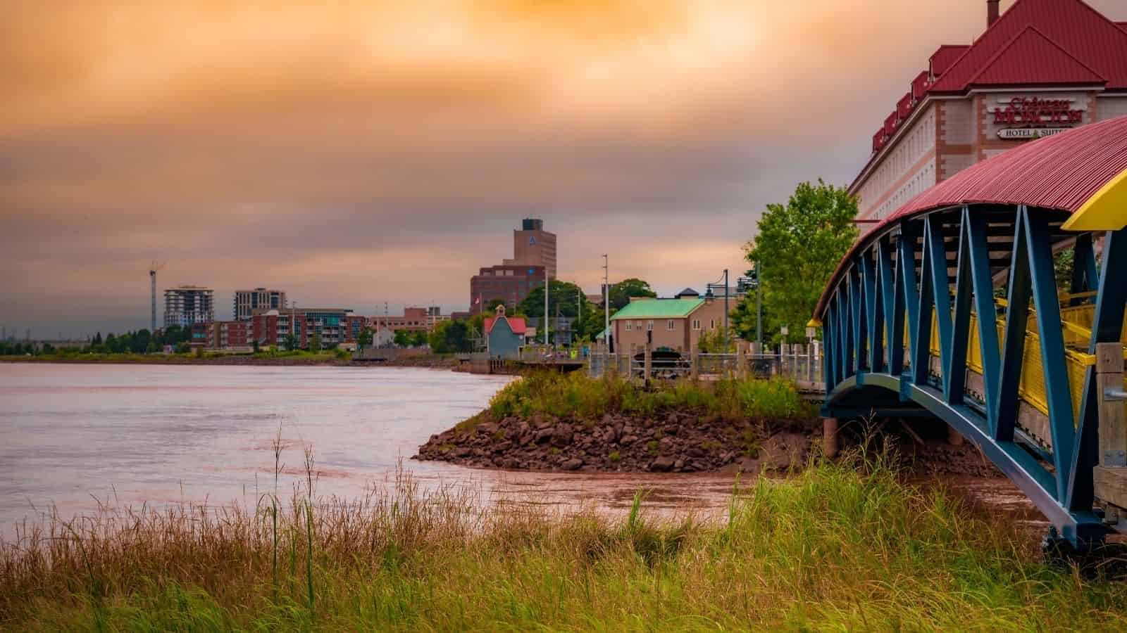 A blue pedestrian bridge crosses a river, leading to a city skyline under an orange, cloudy sky.