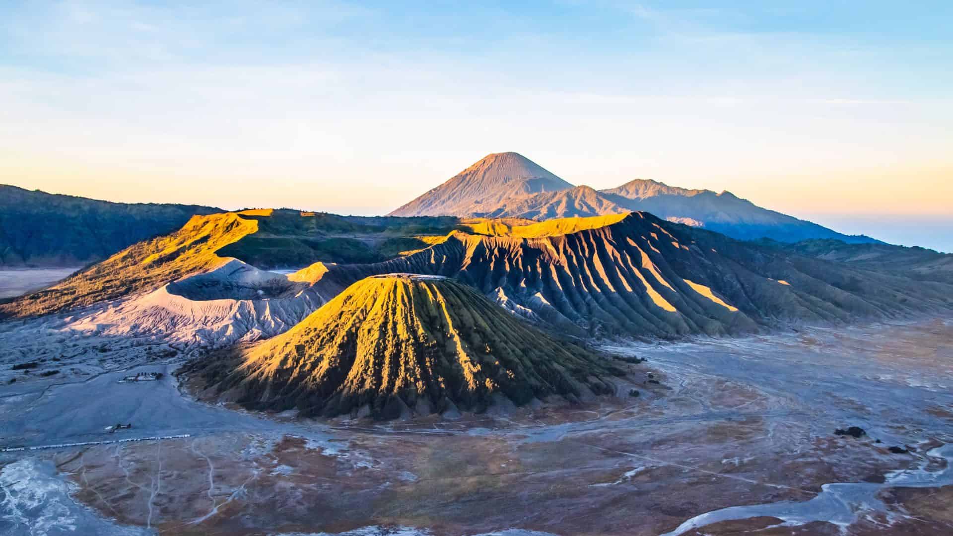 Sunlit volcanic mountains rise above a barren landscape under a clear blue sky at sunrise.