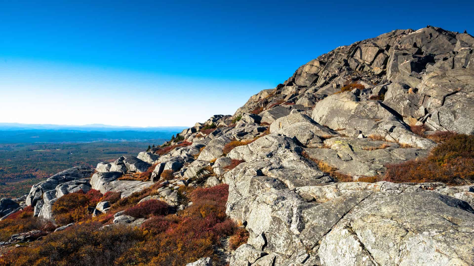 Large rocky mountain slope with scattered shrubs under a clear blue sky and distant landscape views.