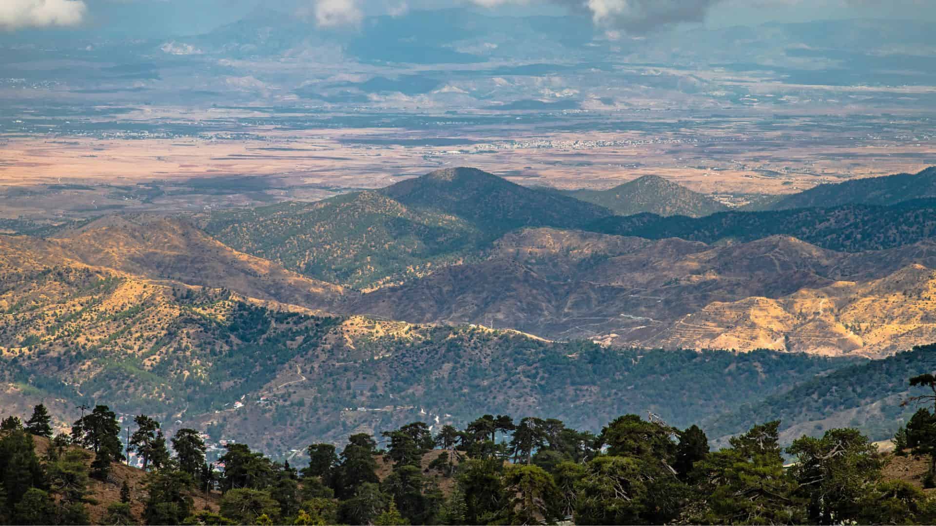 Mountain range with forested hills in the foreground and a vast plain under a partly cloudy sky in the background.