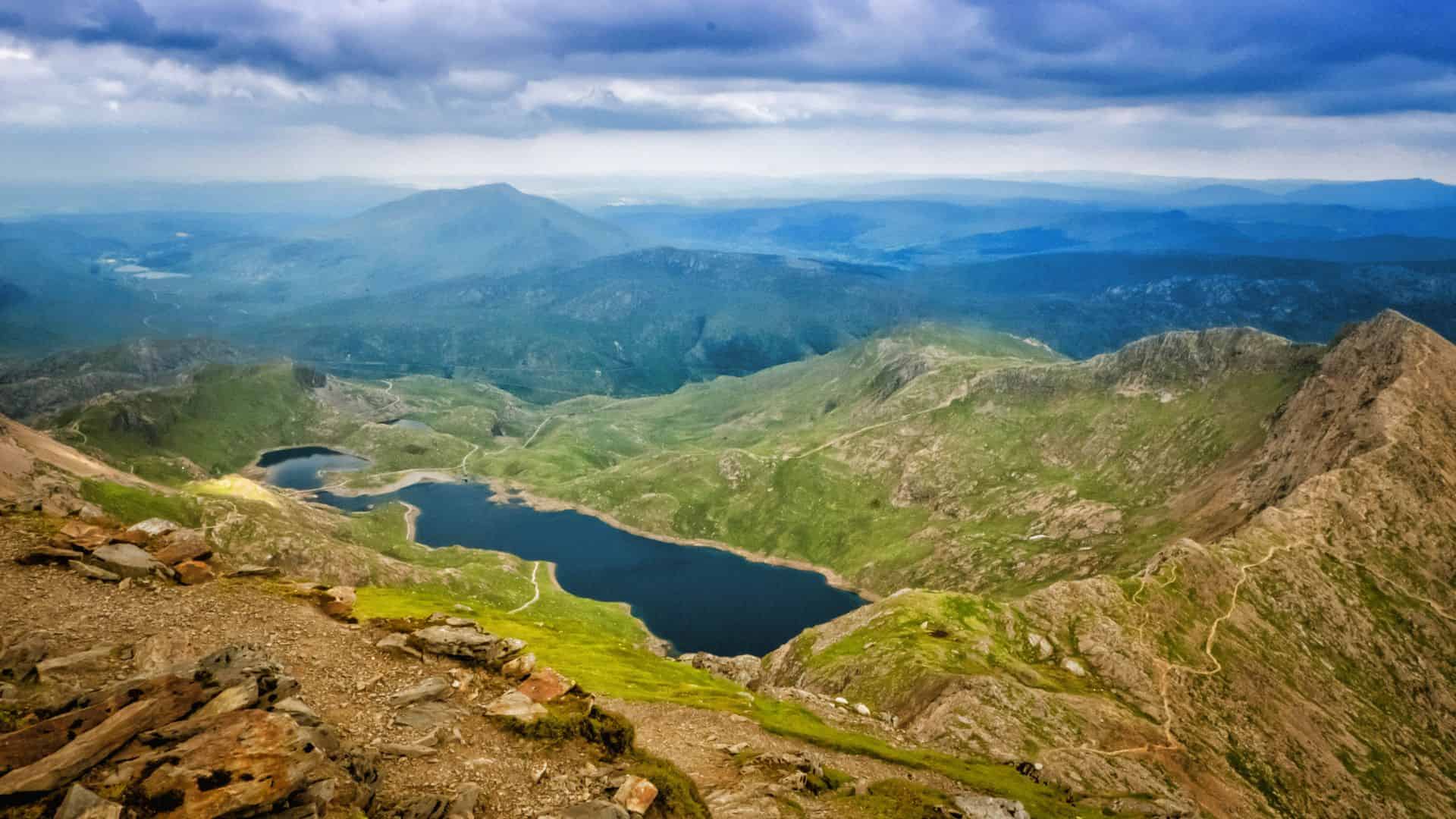 Mountain landscape with rugged peaks, green valleys, and a dark blue lake under a cloudy sky.
